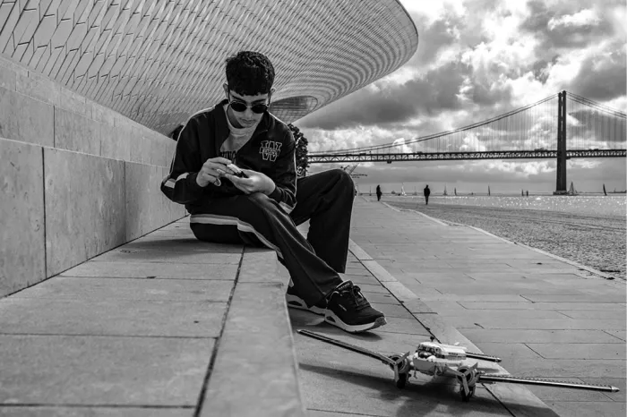 Jongeman met zonnebril zit op de stenen kade onder het golvende dak van het MAAT in Lissabon en kijkt naar zijn telefoon, met een drone naast zich en de brug 25 de Abril aan de horizon.