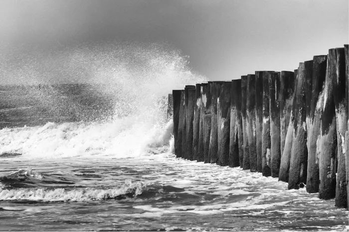 Hoog opspattende golf breekt tegen een rij verweerde houten palen op het strand van Wissant, met schuimend water op de voorgrond.
