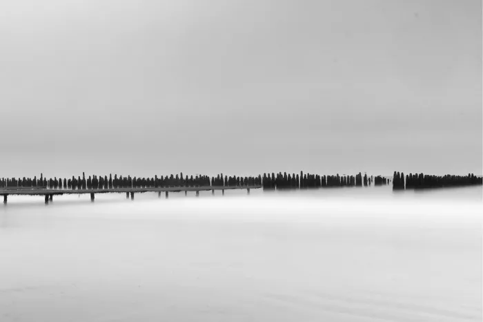 Lange rij lage houten palen trekt op het strand van Wissant een dunne donkere lijn tussen een lichtgrijze lucht en zacht glanzend water.