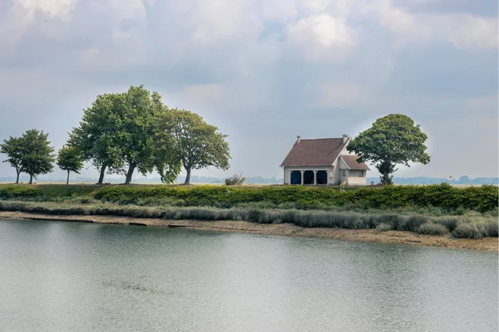 Wit huisje met rood dak tussen weelderige bomen op een groene oever in Honfleur, weerspiegeld in kalm water onder een bewolkte lucht.