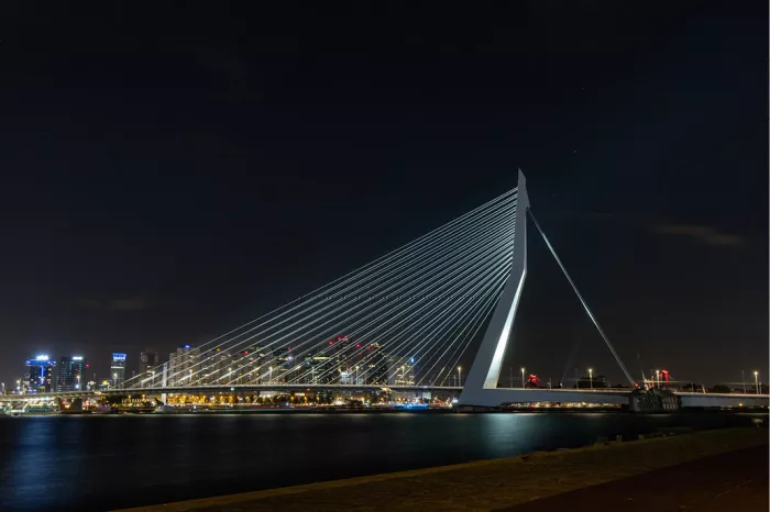 Verlichte Erasmusbrug met haar schuine pyloon en tuikabels tegen een donkere nachthemel, met de skyline van Rotterdam die in het water glinstert.