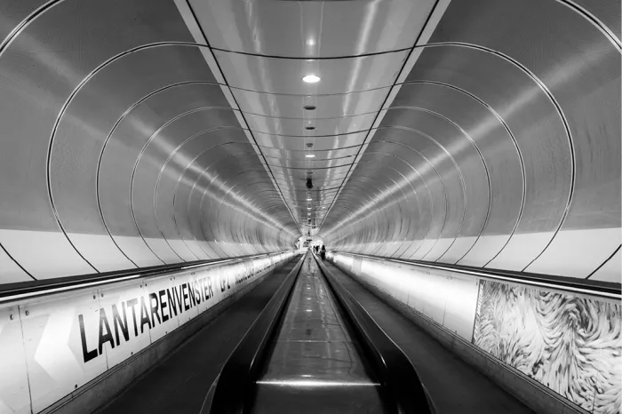 Lange, ronde gang van metrostation Wilhelminaplein in Rotterdam, met rolband en lichtlijnen die in perspectief naar een klein lichtpunt in de verte lopen.