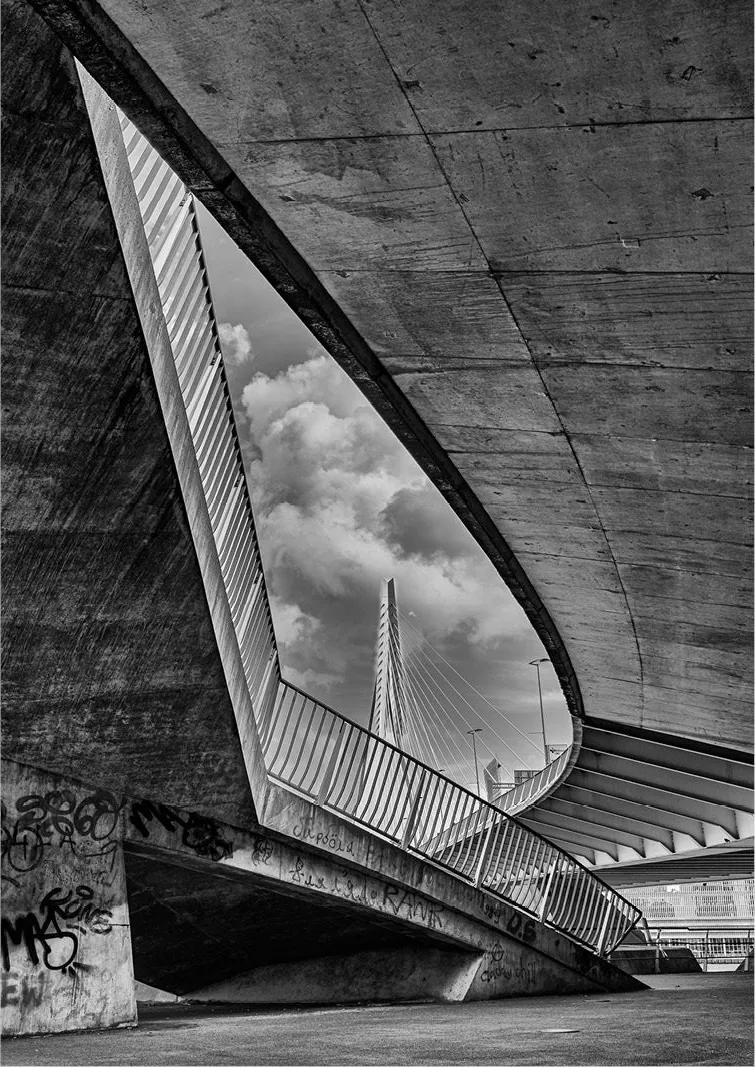 Driehoekige opening in een betonnen constructie kadert de witte pyloon van de Erasmusbrug in Rotterdam en stapelwolken, met graffiti op de voorgrond.