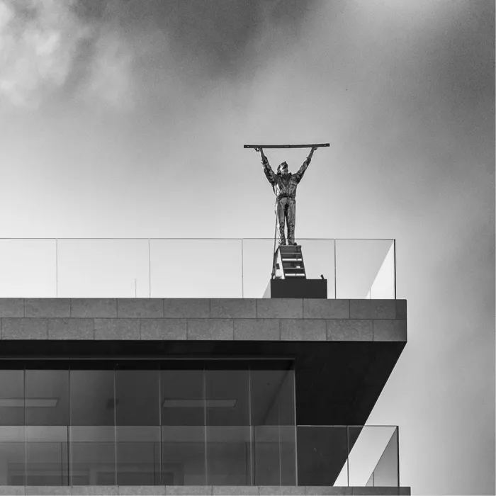 Bronzen beeld Man die de wolken meet op de rand van een appartementsgebouw in Waregem, met een lange staaf boven het hoofd tegen een grijze lucht.