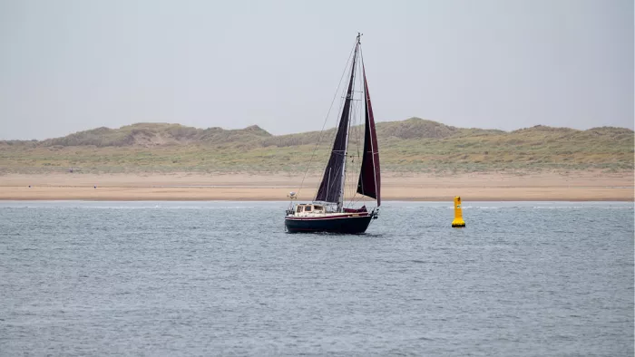 Zeilboot met donkere zeilen naast een gele boei op de zee, met duinen op de achtergrond
