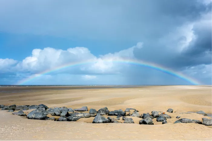 Regenboog boven een breed strand en de zee met grijze stenen op de voorgrond