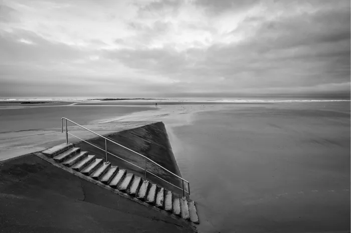 Een minimalistisch zwart-wit beeld van een betonnen trap met metalen leuning die uitkomt op een uitgestrekt, leeg strand bij een bewolkte hemel.