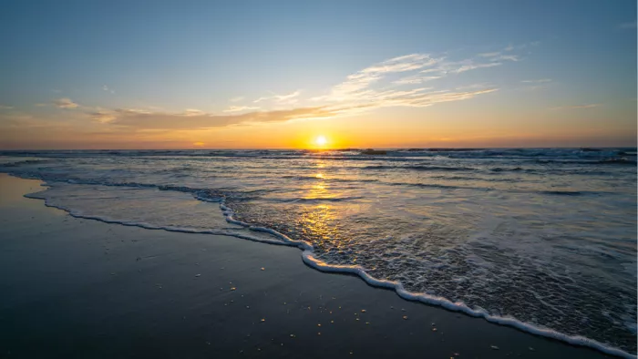 Een prachtige zonsondergang waarbij de zon precies op de horizon rust. De warme stralen weerspiegelen op het natte zand en het schuim van de zachte golven.