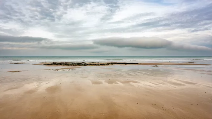 Een weids panorama van een breed strand bij eb. Een opvallende, langgerekte wolk domineert de horizon boven de rustige zee.