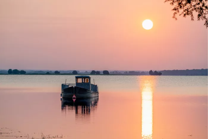 Aangemeerde boot ligt op het water terwijl een gloeiende zon het tafereel in oranje en roze licht zet, met een lange lichtreflectie op het wateroppervlak.