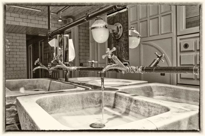 Sepia foto van oude marmeren wastafels met koperen kranen, een dunne waterstraal en zeepdispensers tegen een betegelde muur