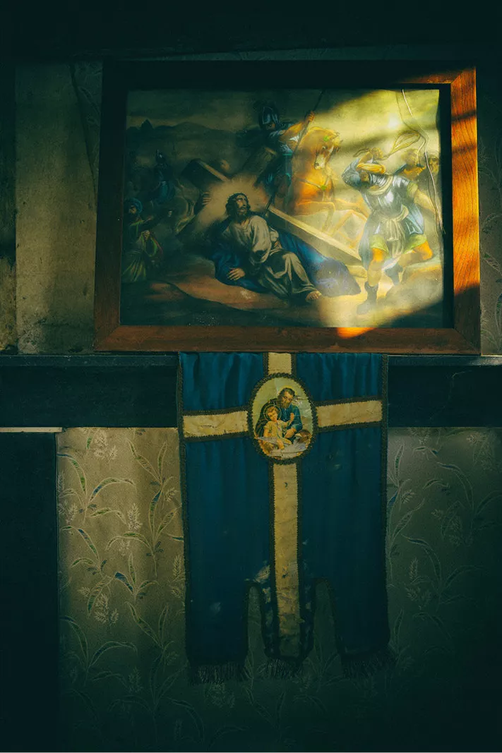 Verlaten huis met achtergebleven religieuze voorwerpen. Het zonlicht speelt op het geweldadige tafereel terwijl Jezus  zijn kruis draagt. Het vredige rust in de schaduw. Aanklacht tegen geweld 