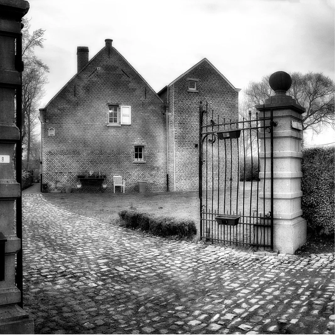 Zwart-wit foto van een bakstenen huis met kasseioprit, stenen poortpijlers en een open smeedijzeren poort