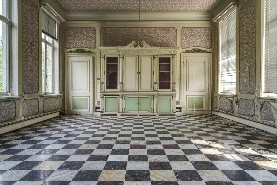 Lege woonkamer in een verlaten herenhuis met dambordvloer, een witte kast met groene panelen en daglicht door hoge ramen