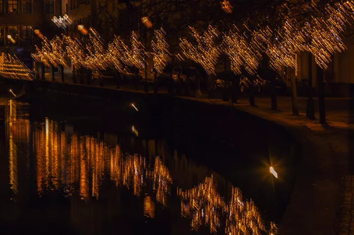 Bomen met gouden kerstlichtjes langs een kaai bij nacht, als bewogen strepen gespiegeld in het water