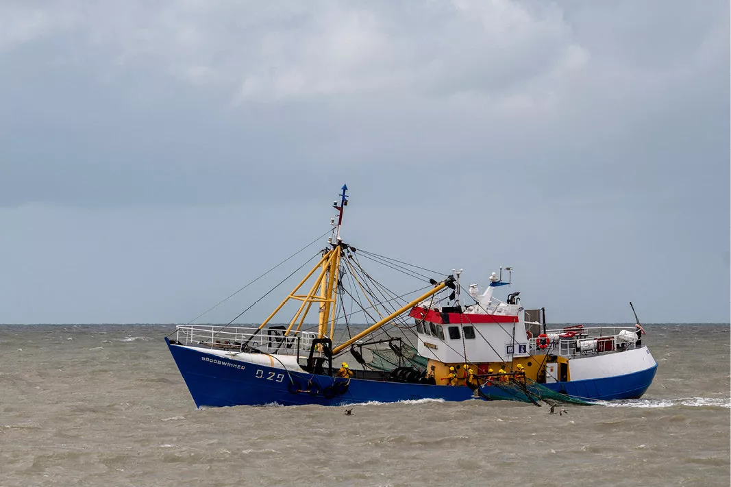 Vissersschip met blauwe romp en gele masten vaart over woelig water onder een bewolkte lucht
