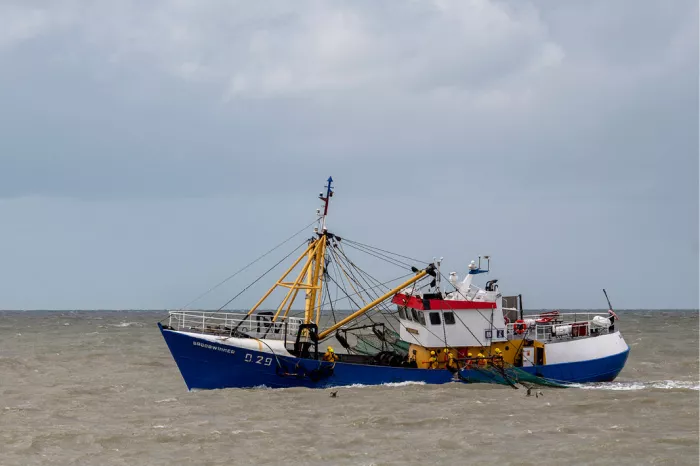 Vissersschip met blauwe romp en gele masten vaart over woelig water onder een bewolkte lucht
