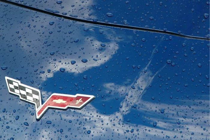 Blauwe motorkap van een Corvette met waterdruppels, het embleem met gekruiste vlaggen en weerspiegelde wolken
