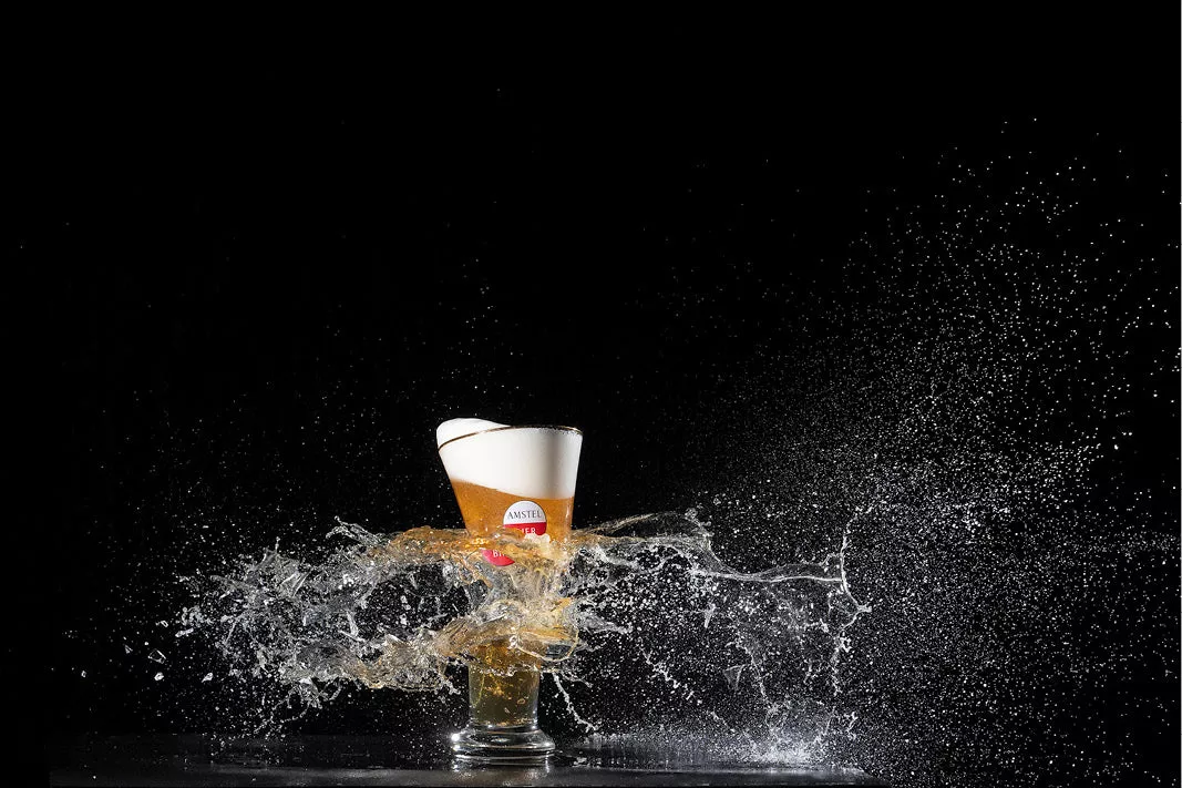 Bierglas wordt door een kogel doorboord, met bier en druppels die opzij spatten tegen een zwarte achtergrond