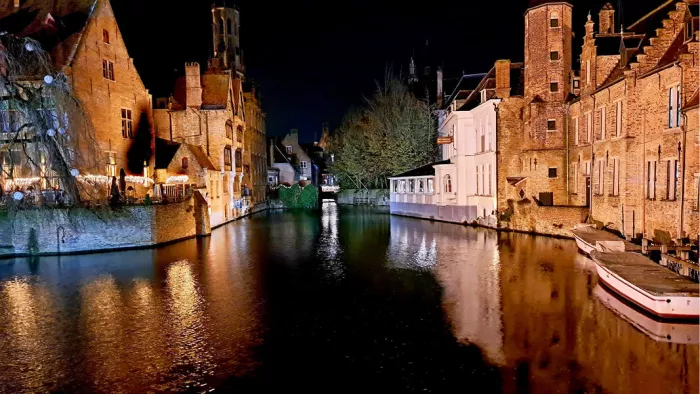 Verlichte gevels en torens van Brugge weerspiegelen 's nachts in de reien, met bootjes aan de kade
