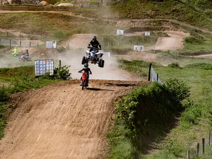 Quad die hoog boven een stoffige crossbaan springt, terwijl een jonge rijder op een rode crossmotor de heuvel op komt.