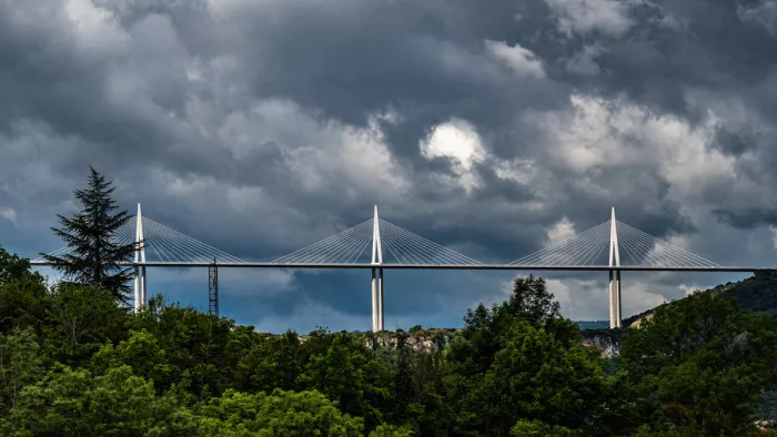 Witte pylonen van een lange tuibrug steken boven een groene heuvelrug uit, onder een zwaar bewolkte lucht met een enkele lichtplek.