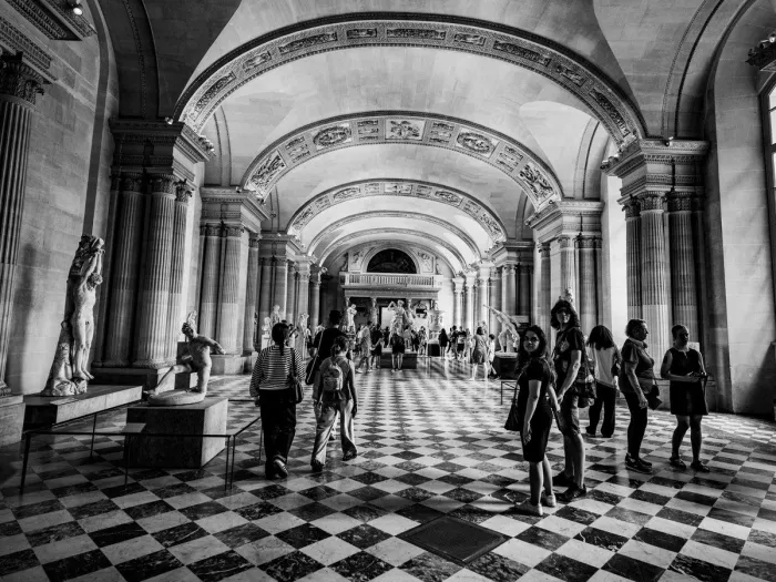 Bezoekers in een museumgalerij met gewelven, beelden en een geblokte vloer, in zwart-wit