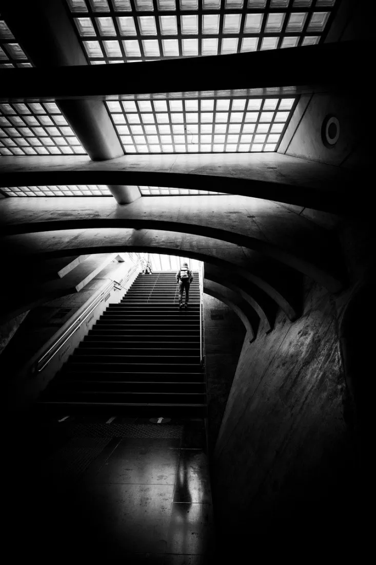 Eenzame figuur beklimt een lange trap onder de betonnen bogen en glazen dakpanelen van station Liège-Guillemins in Luik, in contrastrijk zwart-wit.