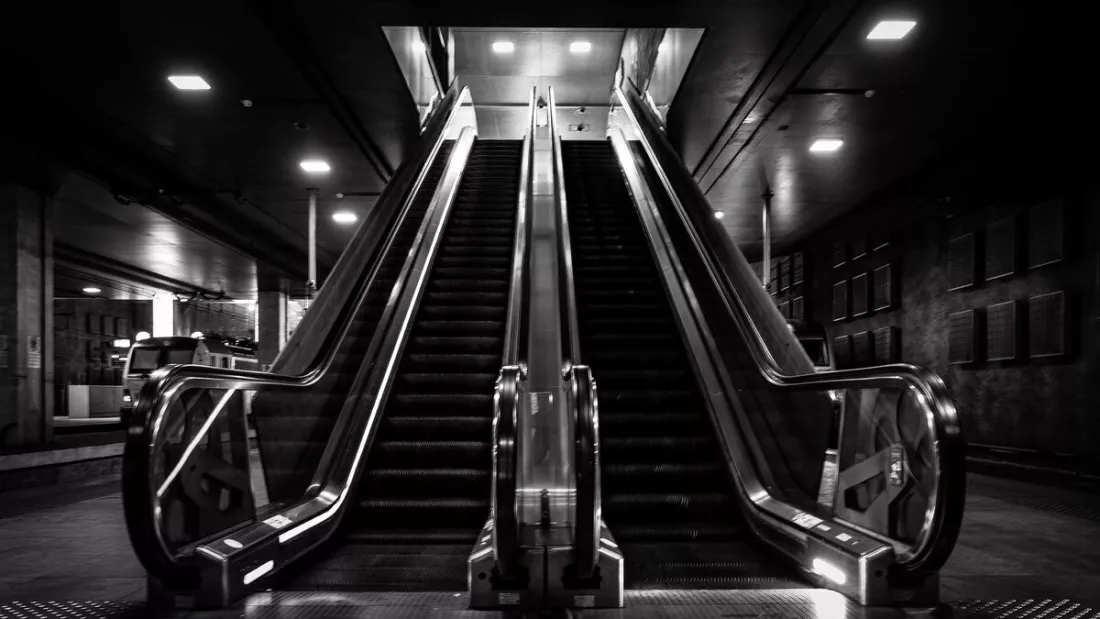 Twee lege roltrappen in een donker station, met glanzende leuningen en vierkante lichtpunten in het plafond.