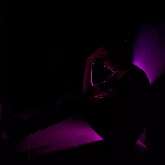 Silhouet van een zittende persoon met de hand aan het hoofd, afgetekend door paars licht op een tegelvloer.
