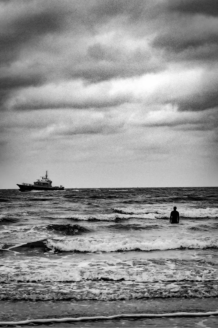 Een eenzame zwemmer trotseert de woelige Noordzee terwijl een patrouilleboot onder een dramatisch wolkendek de horizon doorkruist. Deze sfeervolle kustfoto, vastgelegd in zwart-wit, roept gevoelens op van eenzaamheid, vrijheid en de tijdloze kracht van de zee.