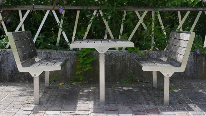 Twee grijze banken met een tafeltje op een terras, voor een witte afsluiting met klimplanten