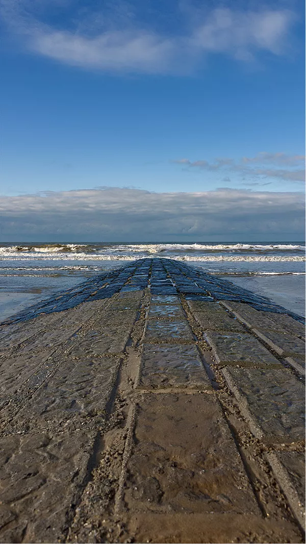 Stenen golfbreker loopt recht de zee in, met brekende golven onder een blauwe lucht