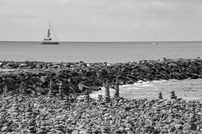 Steenmannetjes van gestapelde stenen op een kiezelstrand met zeilboten op zee, in zwart-wit
