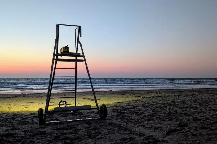 Reddersstoel op wieltjes op het strand na zonsondergang, met geel licht op het zand en een roze lucht