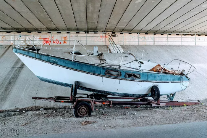 Versleten wit-blauw bootje op een aanhangwagen onder een betonnen brug met graffiti