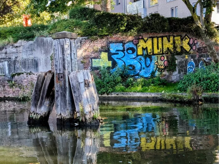 Blauw-gele graffiti op een bakstenen muur langs de Dijle, gespiegeld in het water naast houten palen