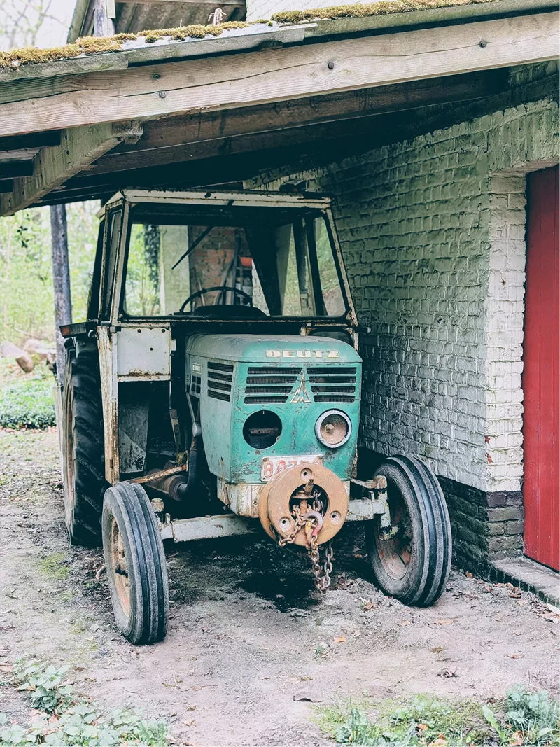 Oude groene tractor onder een houten afdak naast een witte bakstenen muur