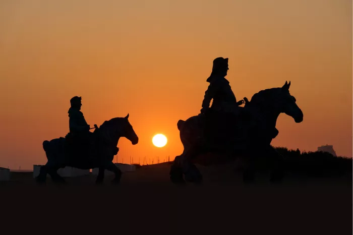 Silhouet van een standbeeld van twee garnaalvissers te paard tegen een oranje lucht met lage zon