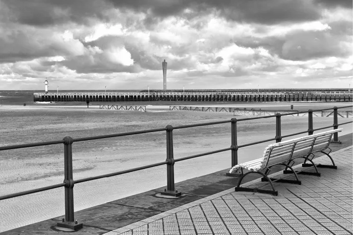 Witte zitbank achter een reling op de zeedijk met het strand en een staketsel onder wolken