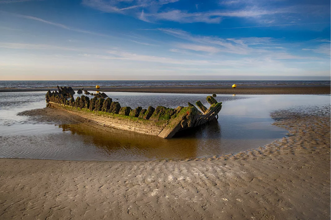 Begroeid scheepswrak in een plas op het strand bij laagtij onder een blauwe lucht