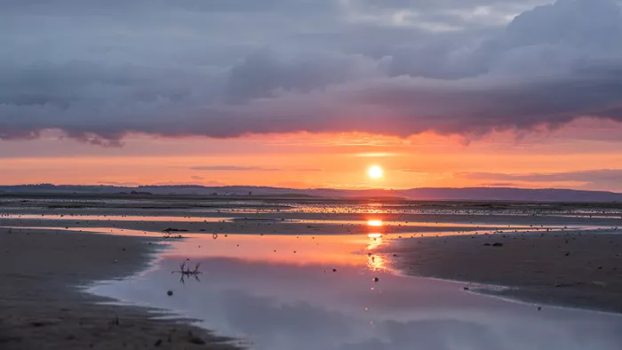 Zon onder een donkere wolkenband boven een nat strand, met een gouden streep in een plas
