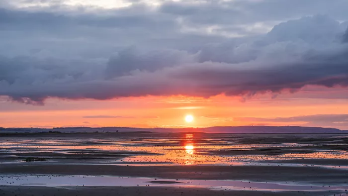 Zonsondergang boven een vlak nat strand met roze en oranje strepen water en een dikke wolkenlaag