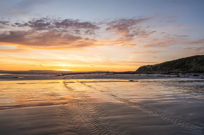 Oranje zonsopgang boven een nat strand met sporen in het zand en een heuvelkam aan zee
