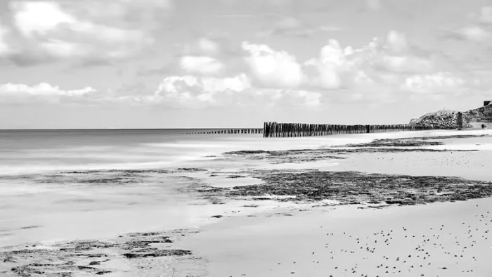 Breed strand met rotsplaten en een verre rij strandpalen onder een wolkenlucht, in zwart-wit