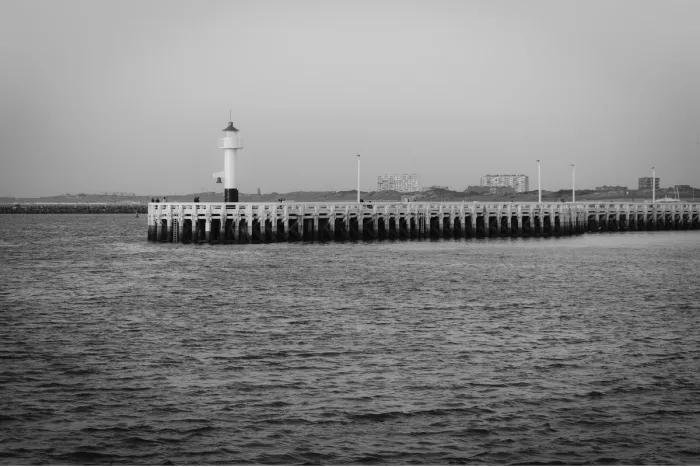 Houten pier met een klein wit vuurtorentje aan het uiteinde, met duinen en gebouwen aan de horizon