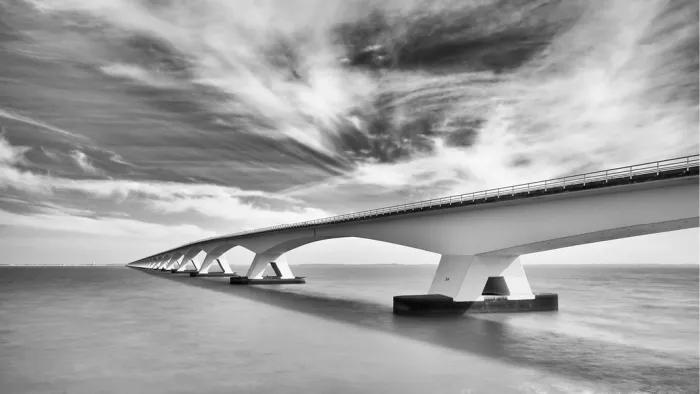 Lange brug met betonnen pijlers over spiegelglad water onder uitwaaierende wolkenstrepen