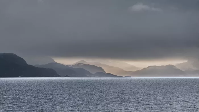 Vervagende bergkammen in tegenlicht boven een grijze zeearm onder een donkere wolkenbank