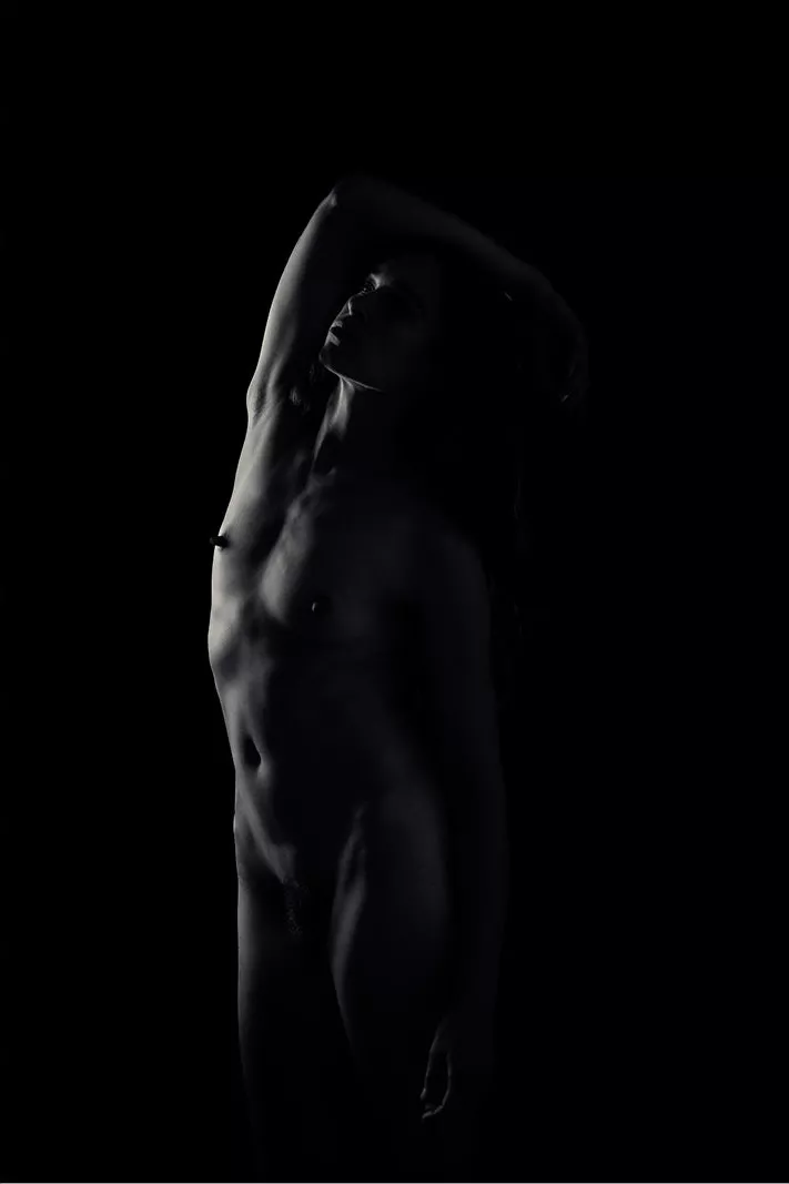 Vrouw met opgeheven gezicht en een arm boven het hoofd, zacht belicht tegen een zwarte achtergrond