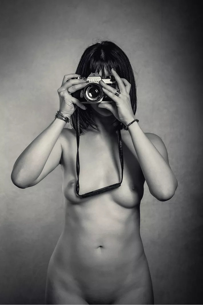 Naakte vrouw fotografeert de kijker met een analoge camera voor het gezicht, tegen een grijze achtergrond